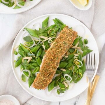 A pistachio crusted salmon filet on a bed of bright green sugar snap peas with a fork.