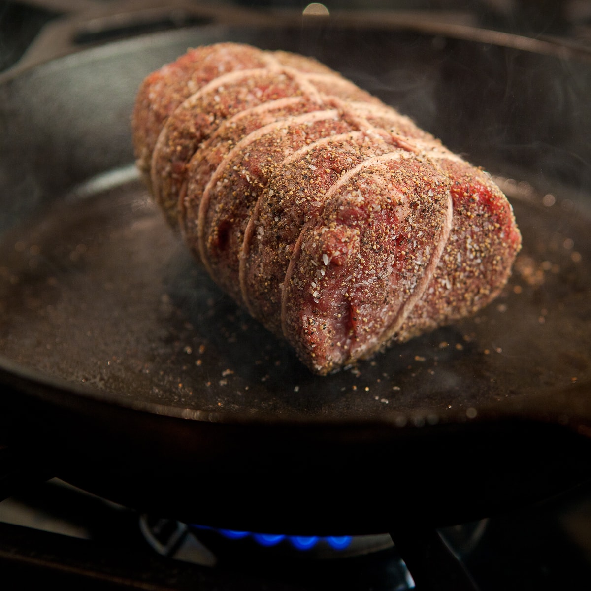 A tied and seasoned beef tenderloin just starting the searing process in a cast iron pan.
