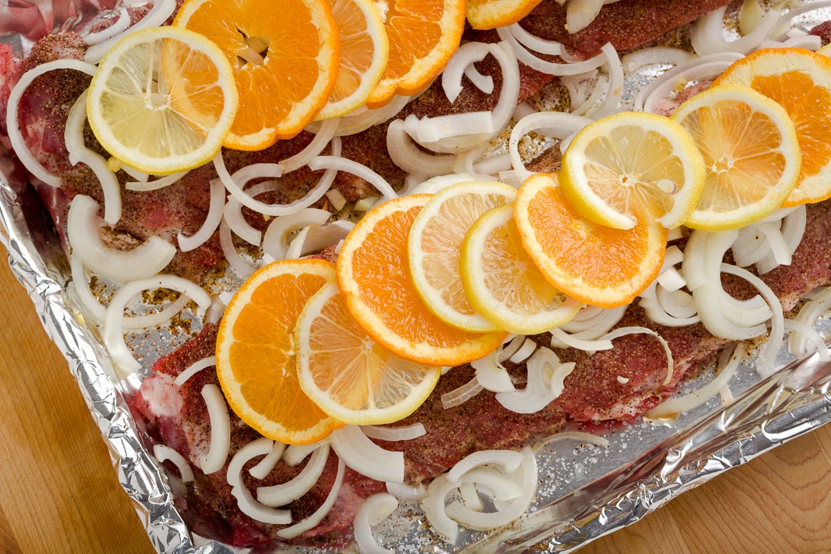 Baby back ribs covered in slice onions, lemons and orange on foil tray.