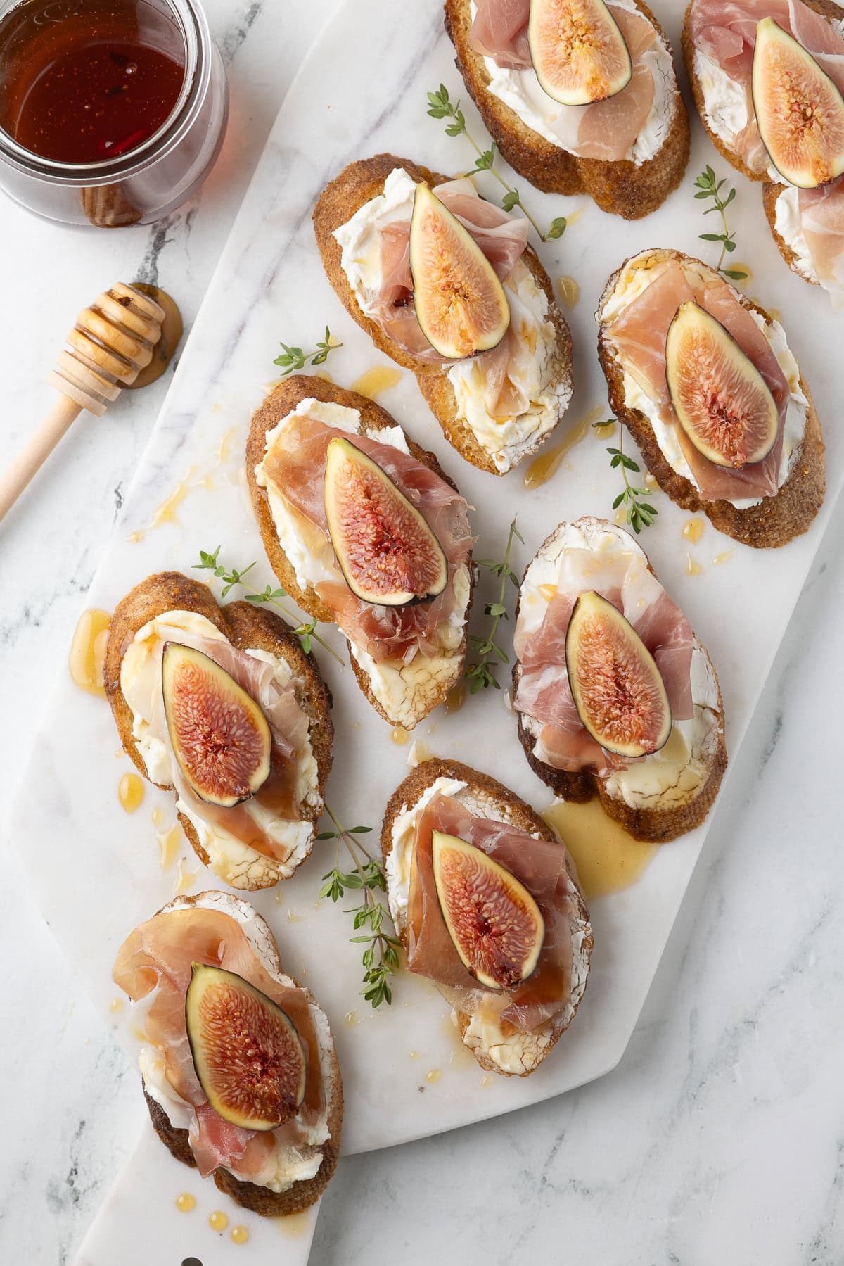 Goat cheese crostini with figs and prosciutto on a marble board with hot honey on the side.