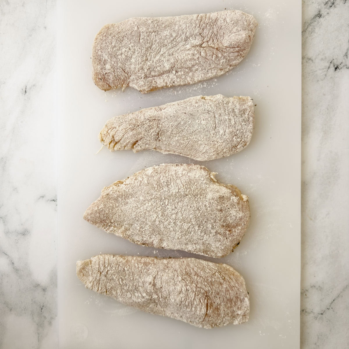 Floured and seasoned chicken breasts on a white cutting board ready for cooking.