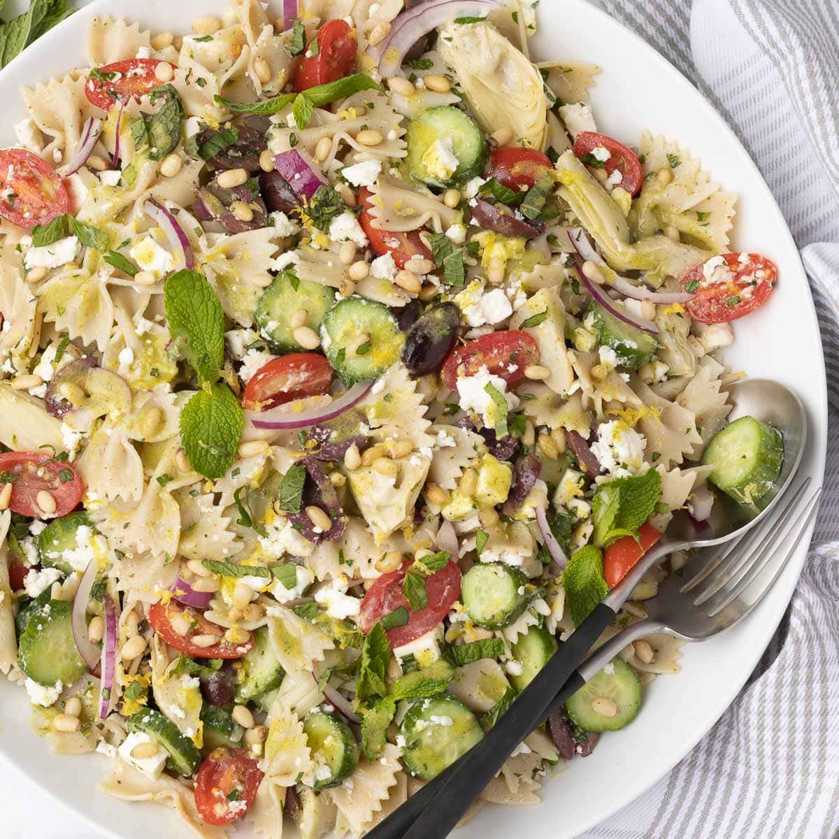 White bowl of colorful farfalle pasta salad with serving utensils.