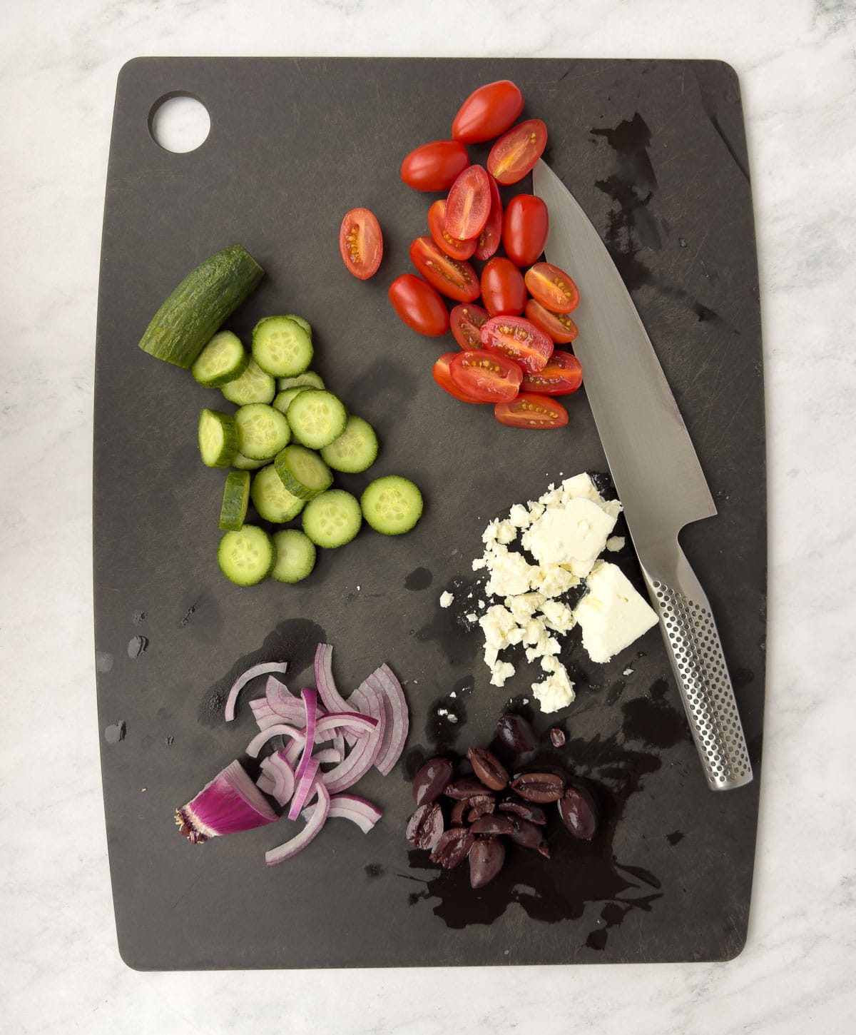 Prepping veggies for pasta salad: tomatoes, olive, cucumbers, feta cheese on a black cutting board.
