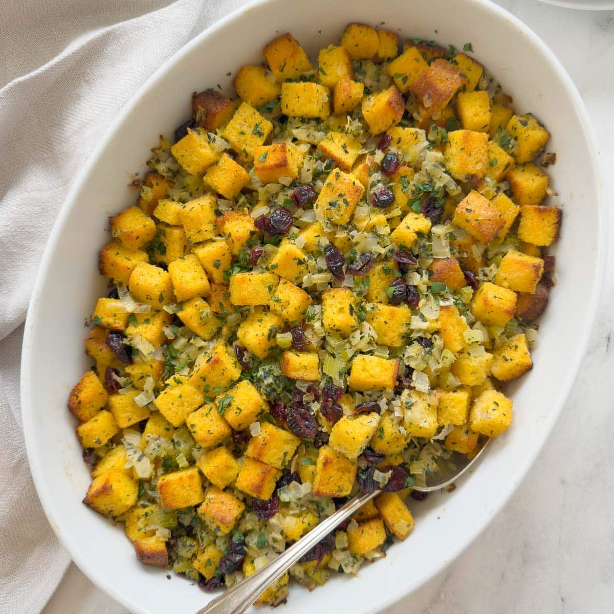 An oval baking dish of golden cornbread dressing with cranberries and veggies, and a silver serving spoon.