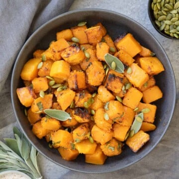 A gray bowl of air fryer butternut sqhuas with pepitas and sage leaves with gray napkin.