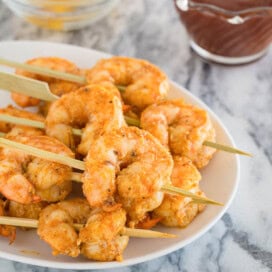 A small appetizer plate of golden baked shrimp on wooden skewers with cocktail sauce.