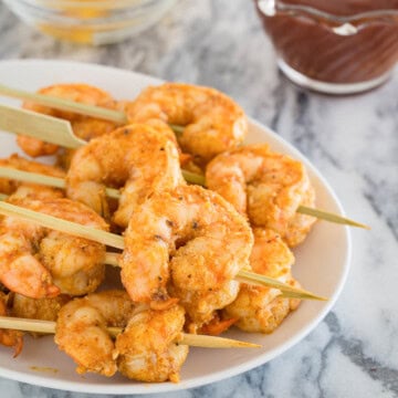 A small appetizer plate of golden baked shrimp on wooden skewers with cocktail sauce.