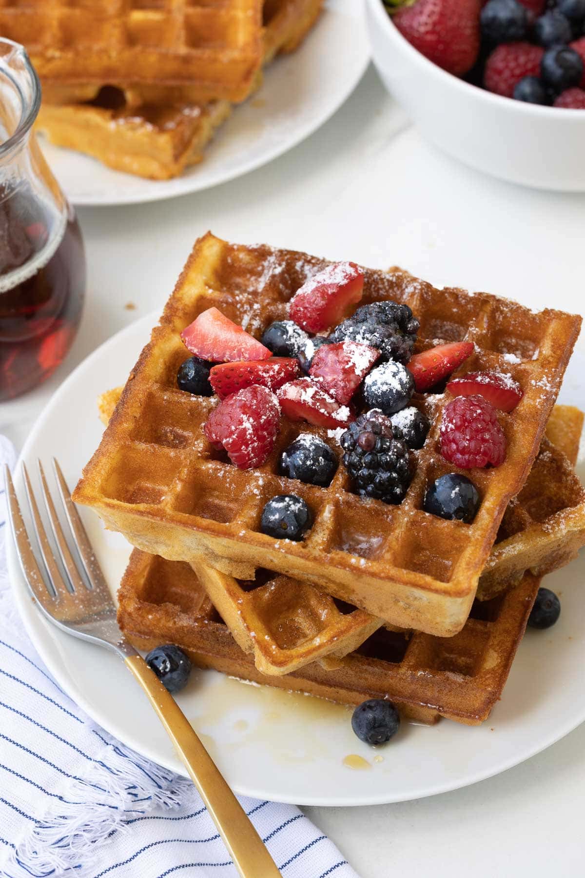A stack of golden crispy gluten-free waffles with berries, maple and powdered sugar.