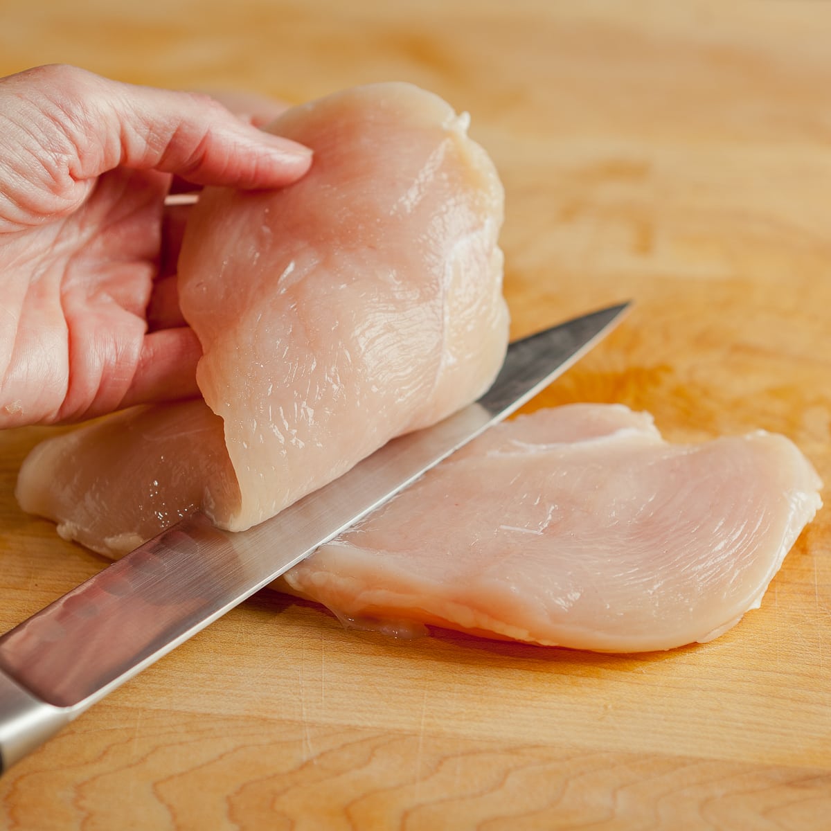 A chicken breast being sliced into half for a cutlet with a thin sharp knife.
