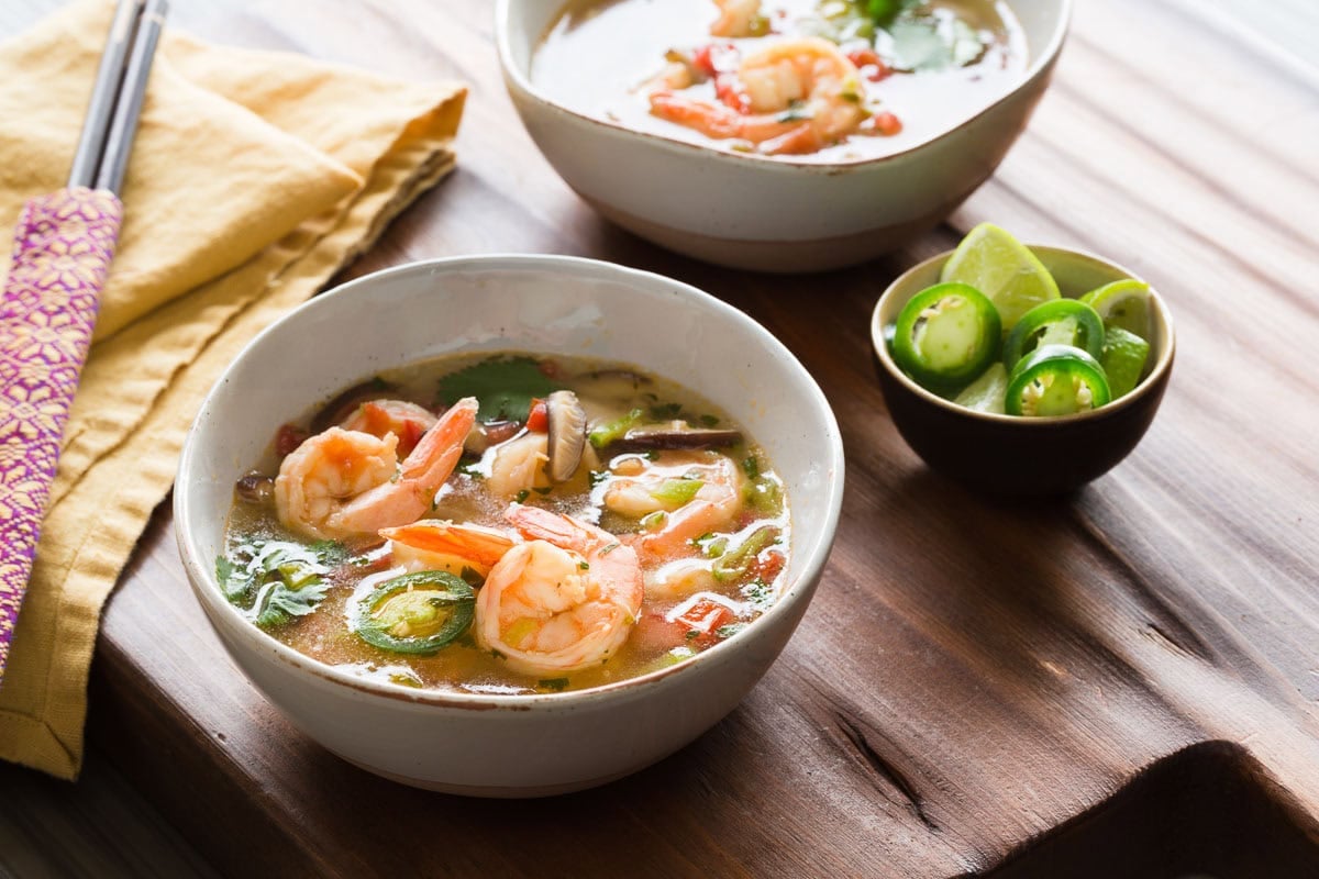 Two bowls of Thai shrimp soup on a wooden table with a small bowl of lime wedges.