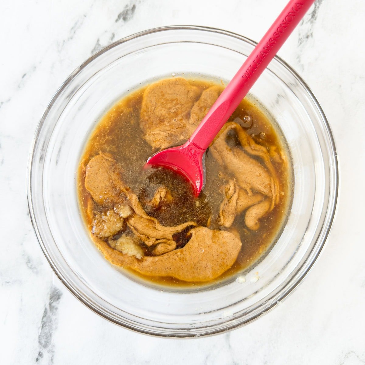 Almond butter and ingredients for Asian dipping sauce in a glass bowl before blending with a red spatula. 