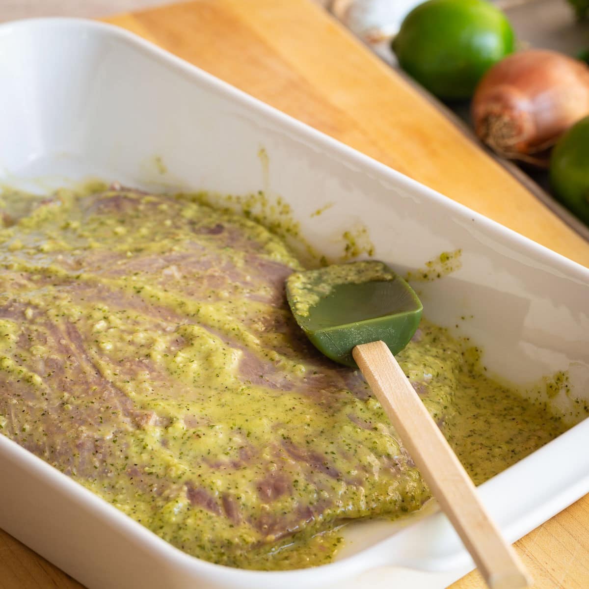 Flank steak marinating in a white casserole dish with a spatula.
