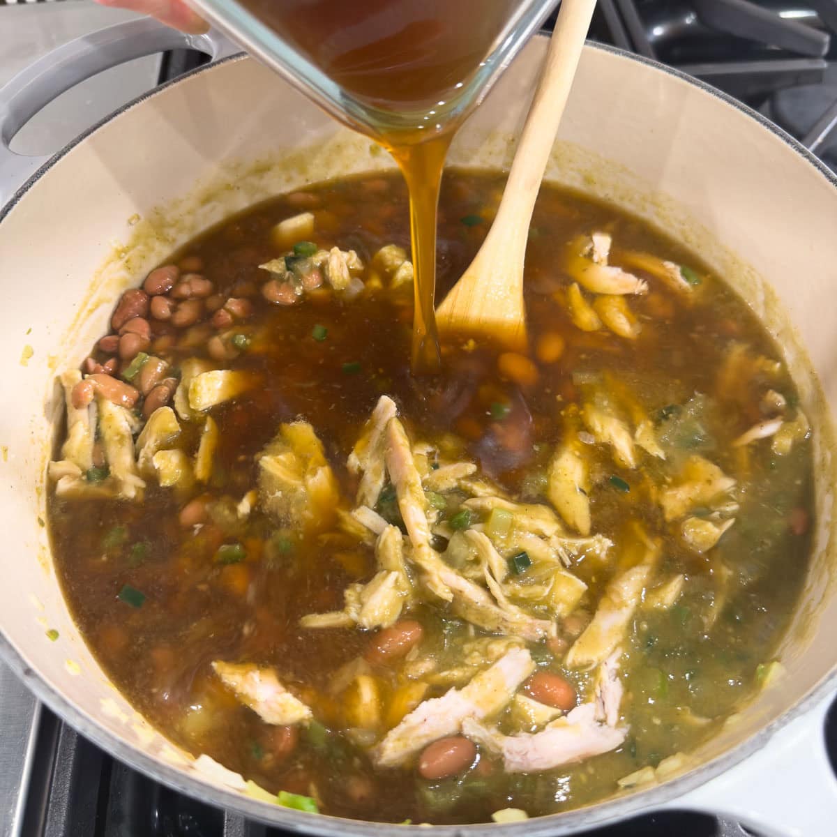 Deep brown turkey broth pouring into a soup pot of soup with a wooden spoon in the back.