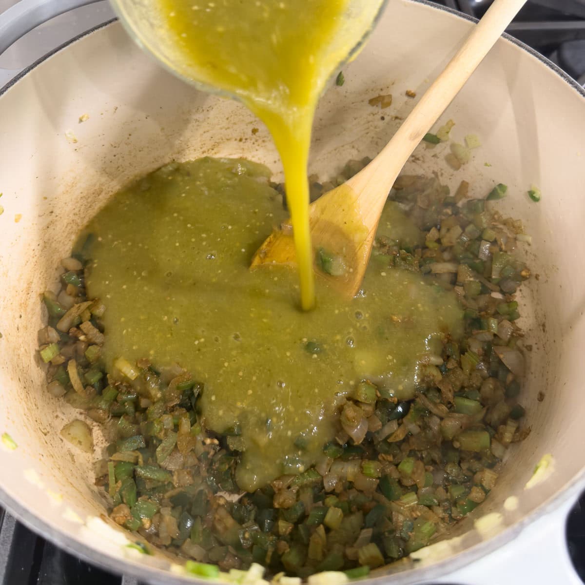Adding tomatillo sauce to a pot of sautéd vegetables, with a wooden spoon.