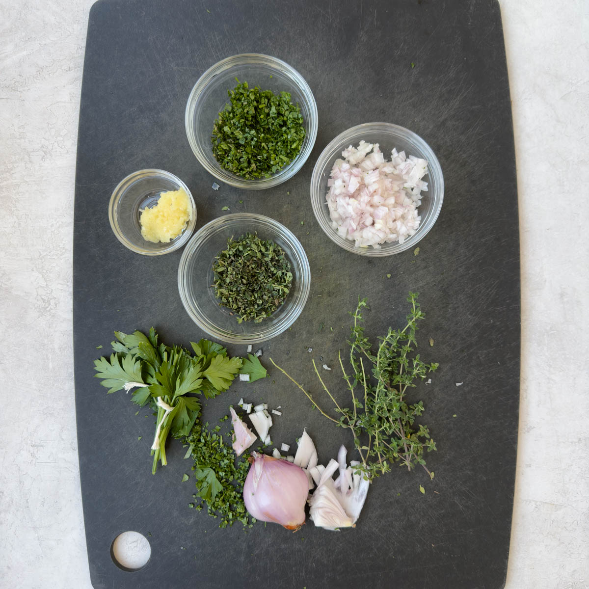 A black cutting board with chopped herbs, shallots and garlic.