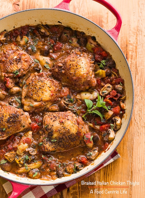 Braised Italian chicken thighs in a shallow pan with vegetables.