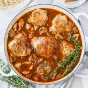 A round pan of golden chicken thighs in red sauce with rosemary on the side.