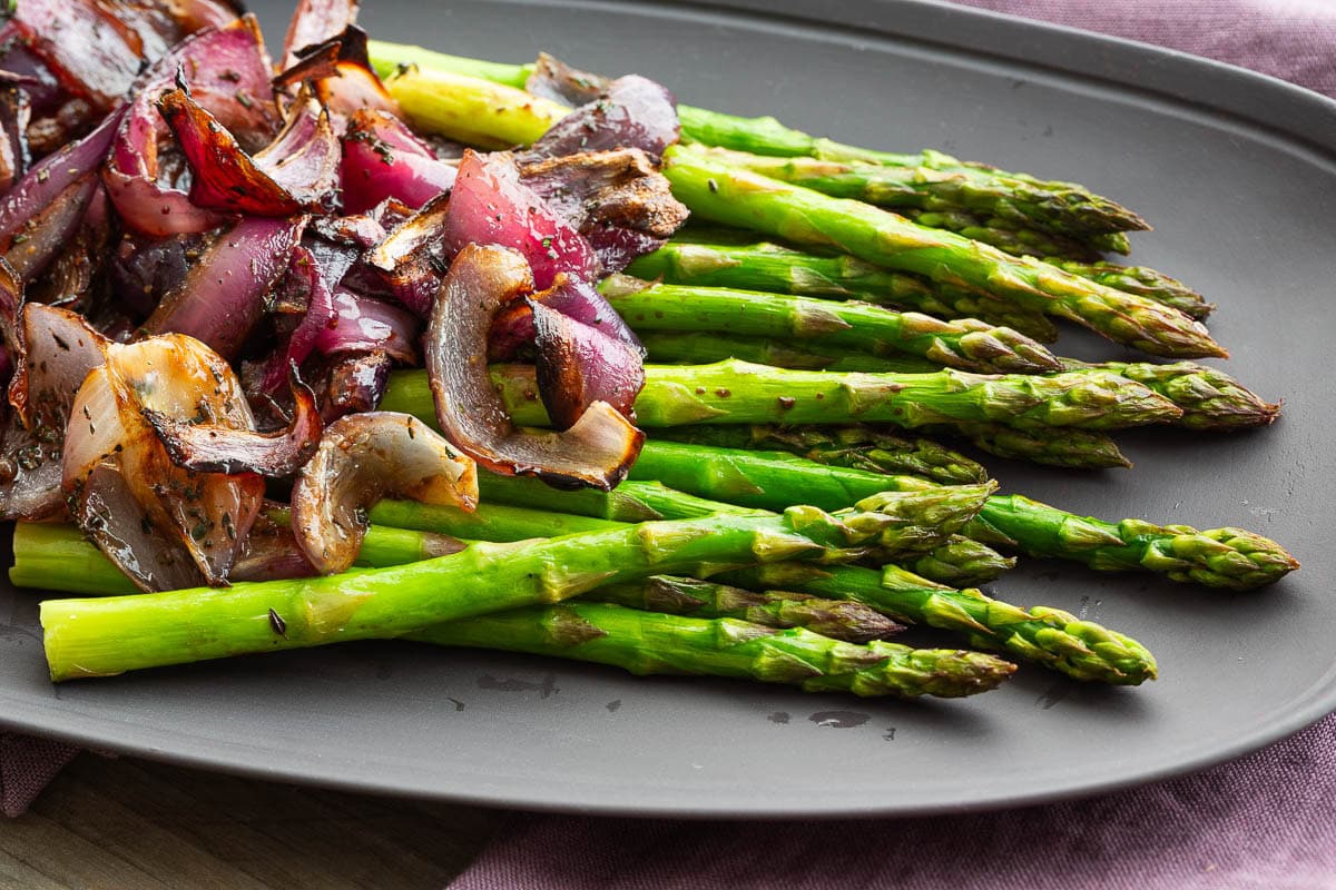 Roasted red onions piled on green asparagus spears on a gray platter.