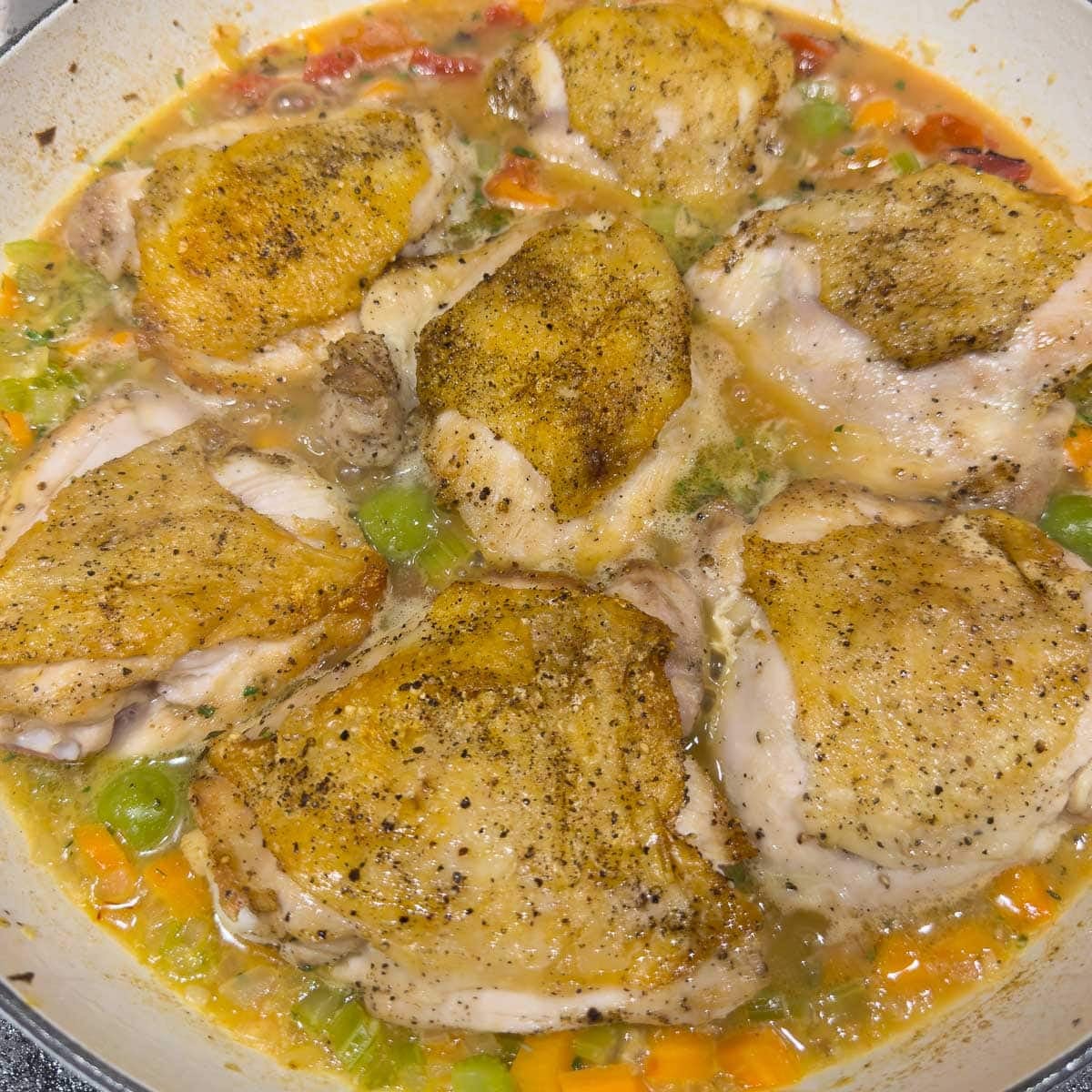 Browned chicken thighs in a wide pan with vegetables and broth
