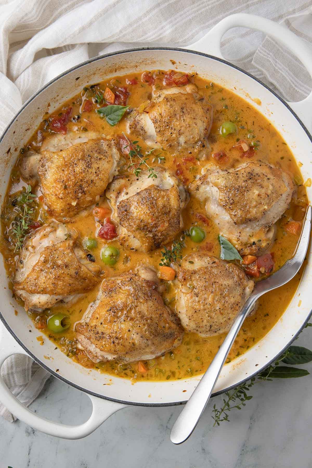 A round white pan of mediterranean chicken thighs with sauce, vegetables, and olives. 