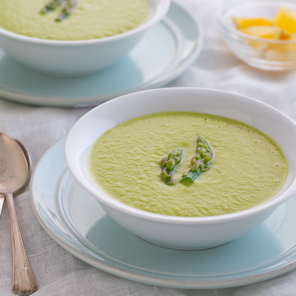 Bright green asparagus soup in a white bowl on a bloue plate on the dinner table.