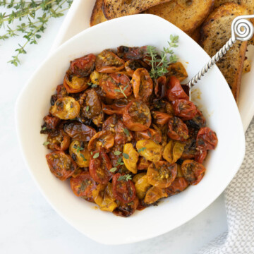 A white bowl of red and gold roasted cherry tomatoes with fresh thyme leaves.