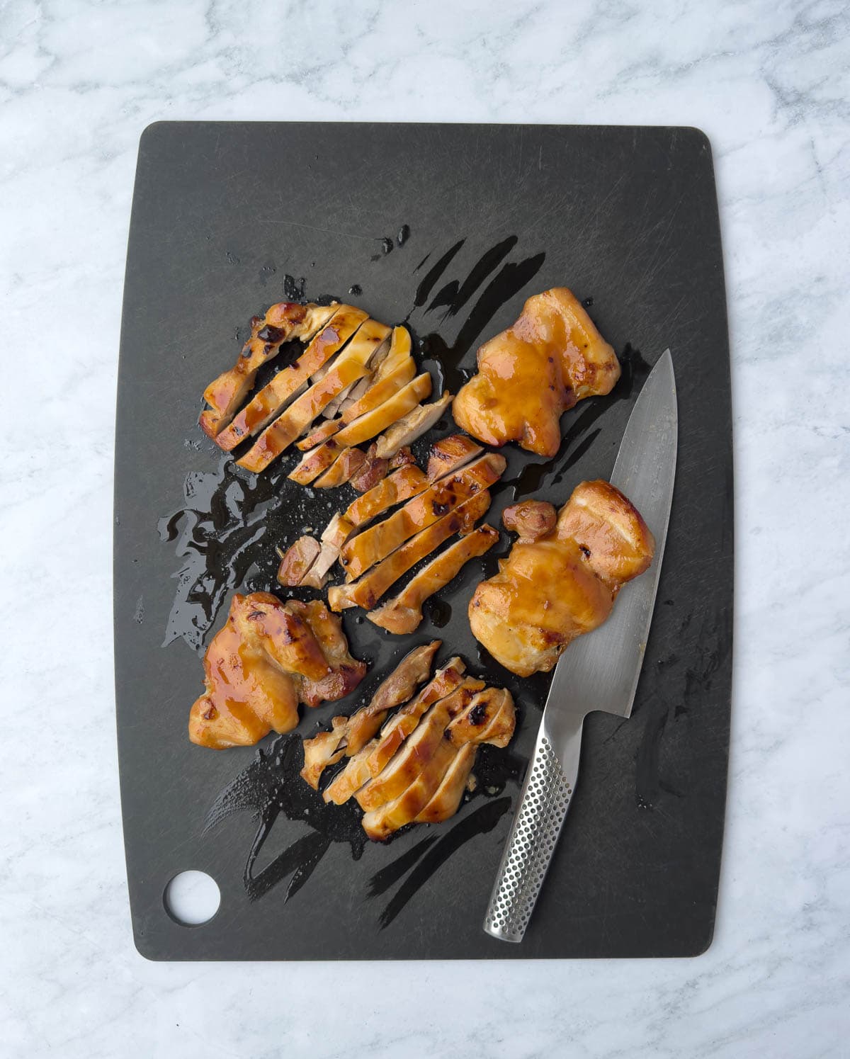 Slicing golden baked chicken thighs on a black cutting board with a chef's knife.