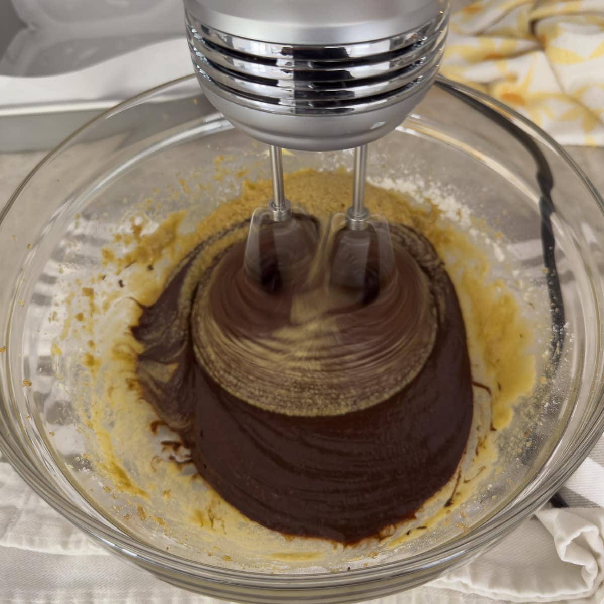 Mixing chocolate brownie batter with an electric hand mixer in a glass bowl.