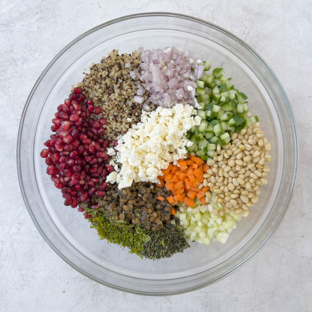 A colorful mix of lentils, quinoa, carrots, pomegranate seeds, herbs and ore for a salad in a glass mixing bowl.