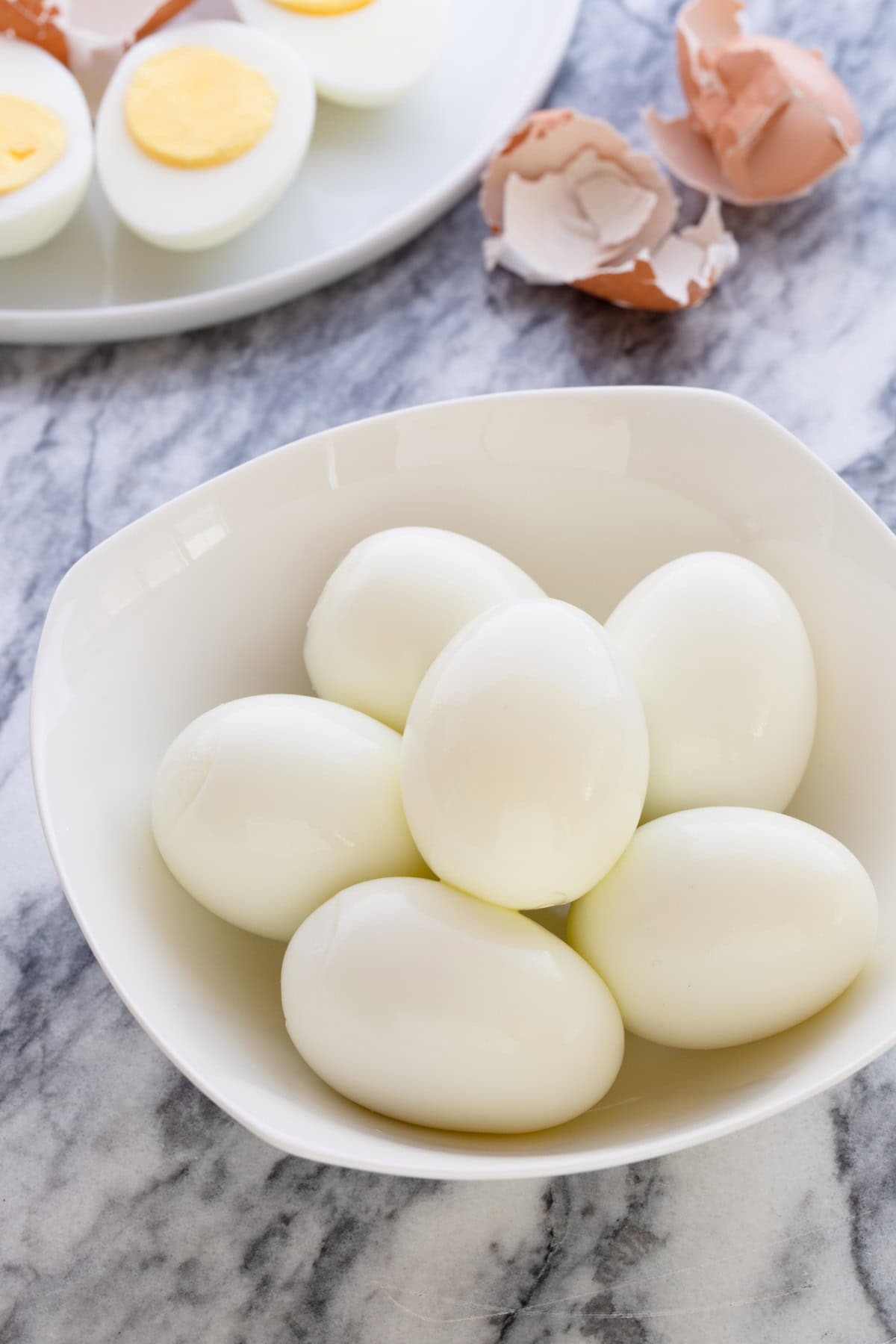 A bowl of peeled perfectly clean hard boiled eggs with peeled and sliced eggs behind with shells.