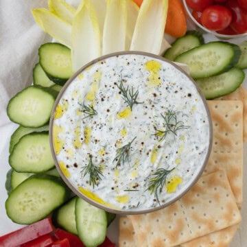 A bowl of cream Greek Tzatziki with dill and olive oil drizzle with raw veggies and crackers.
