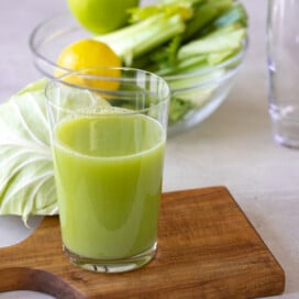 A glass of bright green cabbage juice on a brown cutting board with vegetable bowl behind.