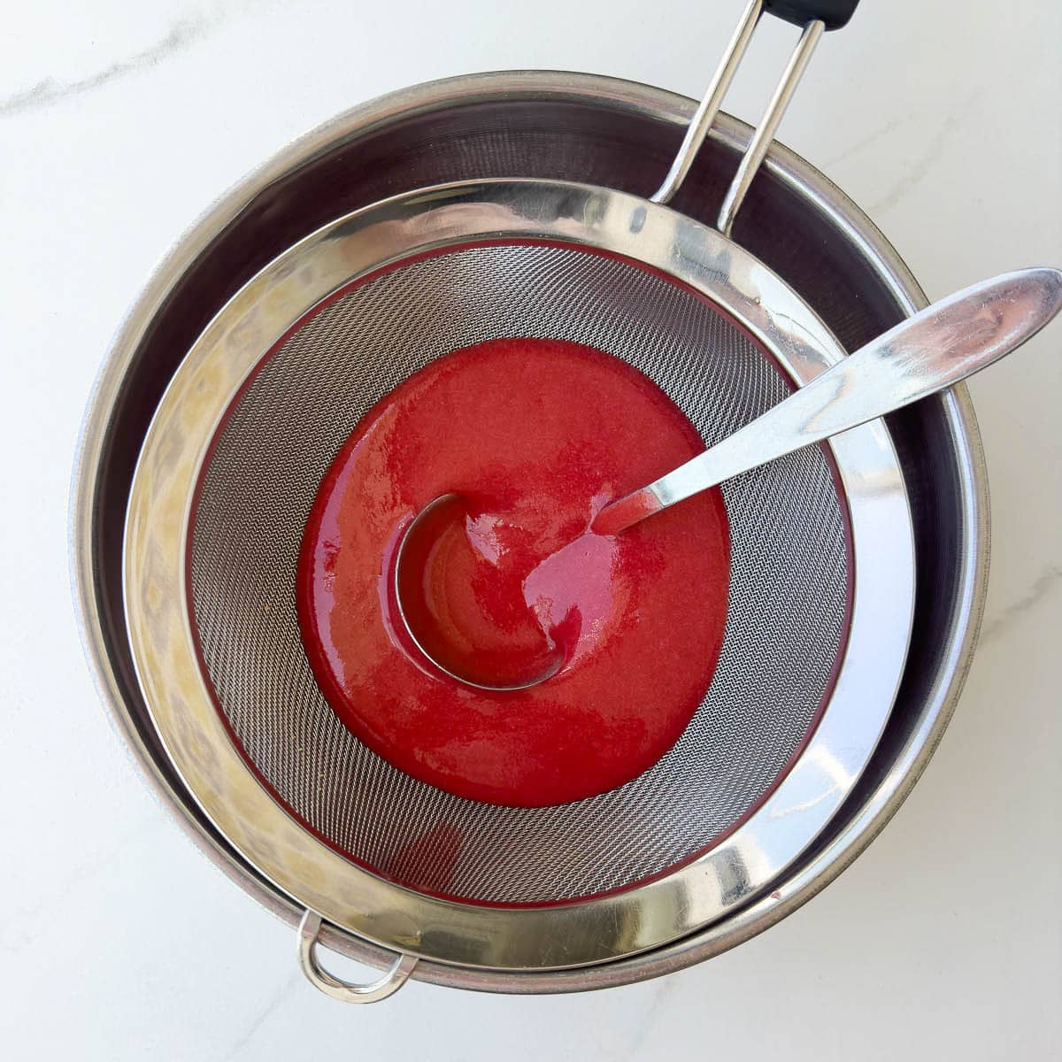Straining strawberry puree through a fine sieve to remove seeds with a ladle.