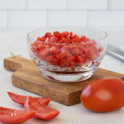 A glass bowl of perfectly diced tomatoes on a cutting board.