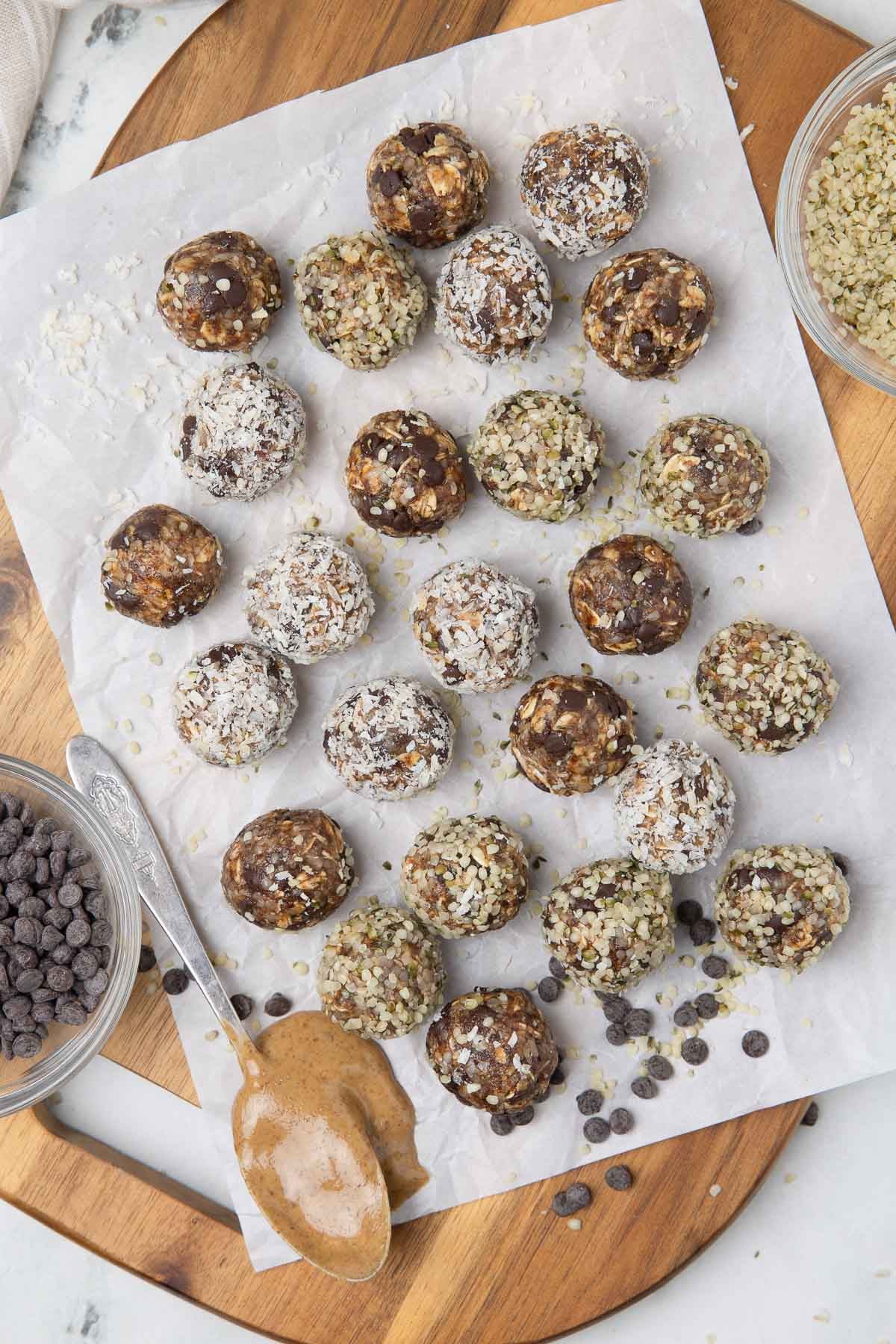 Three types of peanut butter protein balls on a cutting board with chocolate chips, peanut butter, and hemp seeds.