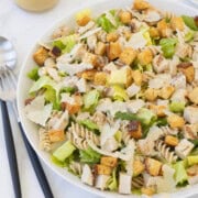 A Caesar pasta salad ready to serve with salad servers and dressing in a marble counter.