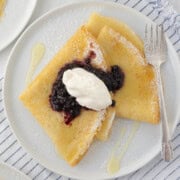 Two golden folded crepes on a white plate with blueberry jam and whipped cream.