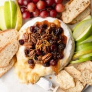 A round wheel of baked brie cheese with a fruit and nut topping, with apples, pears, and crostini.