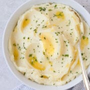 A bowl of creamy mashed potatoes with melted butter and fresh chopped parsley with a silver serving spoon.