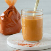A glass jar of bright orange paprika sauce with a golden spoon and a bag of paprika behind.
