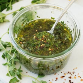 A jar of bright green herb sauce called chimichurri, surrounded by herbs.