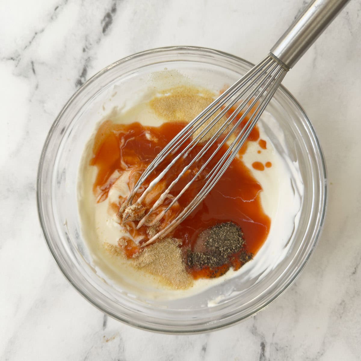 Blending ingredients for buffalo ranch dressing in a glass bowl with a whisk.