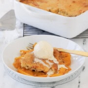 A white bowl of golden peach cobbler with vanilla bean ice cream scoop.