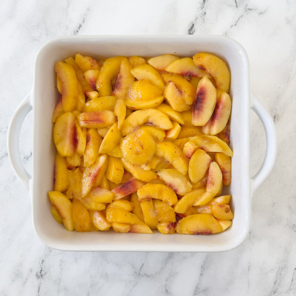 A square baking dish filled with sliced peaches to make ap each cobbler.