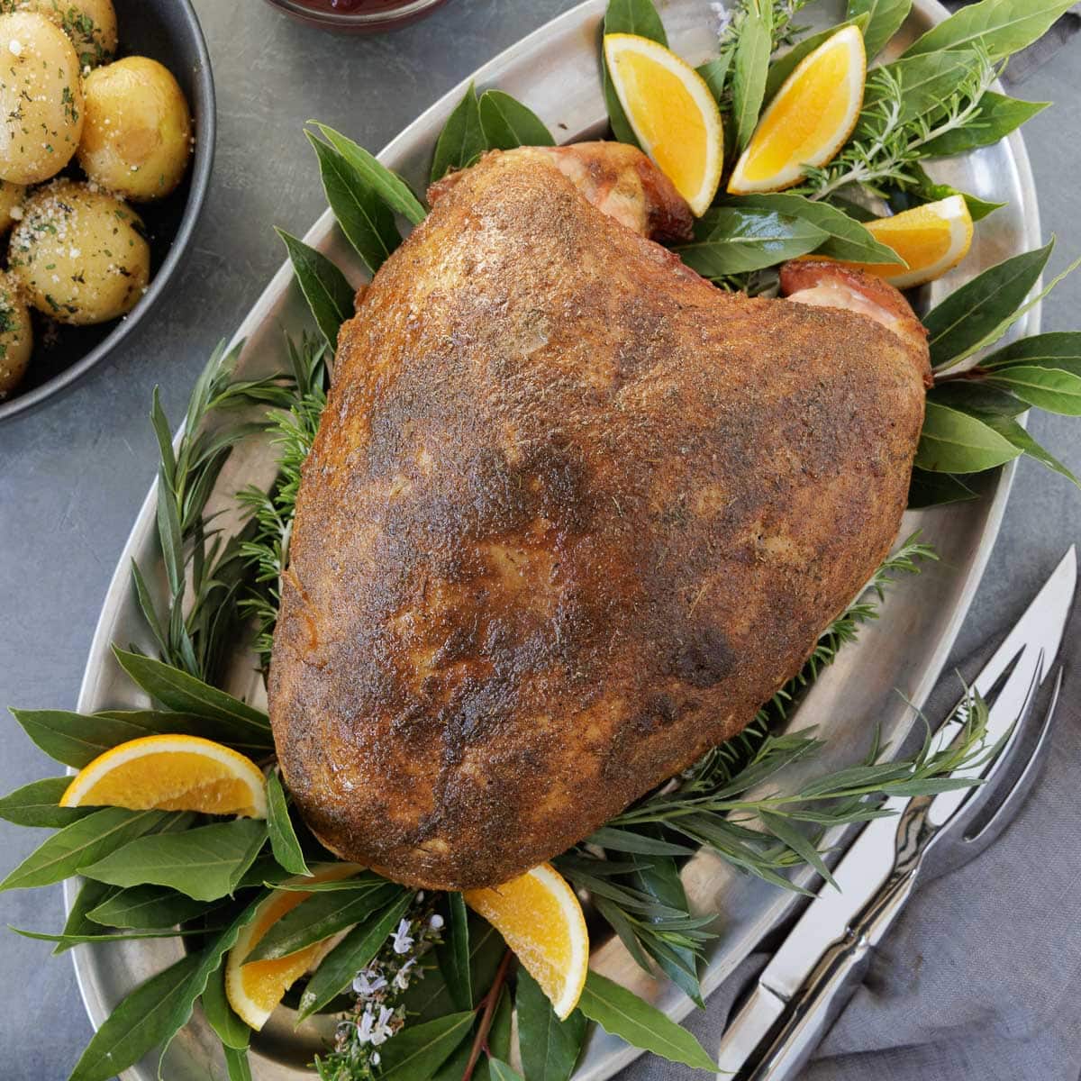 A browned, whole smoked turkey breast in a silver platter with bay leaves, potatoes on side,