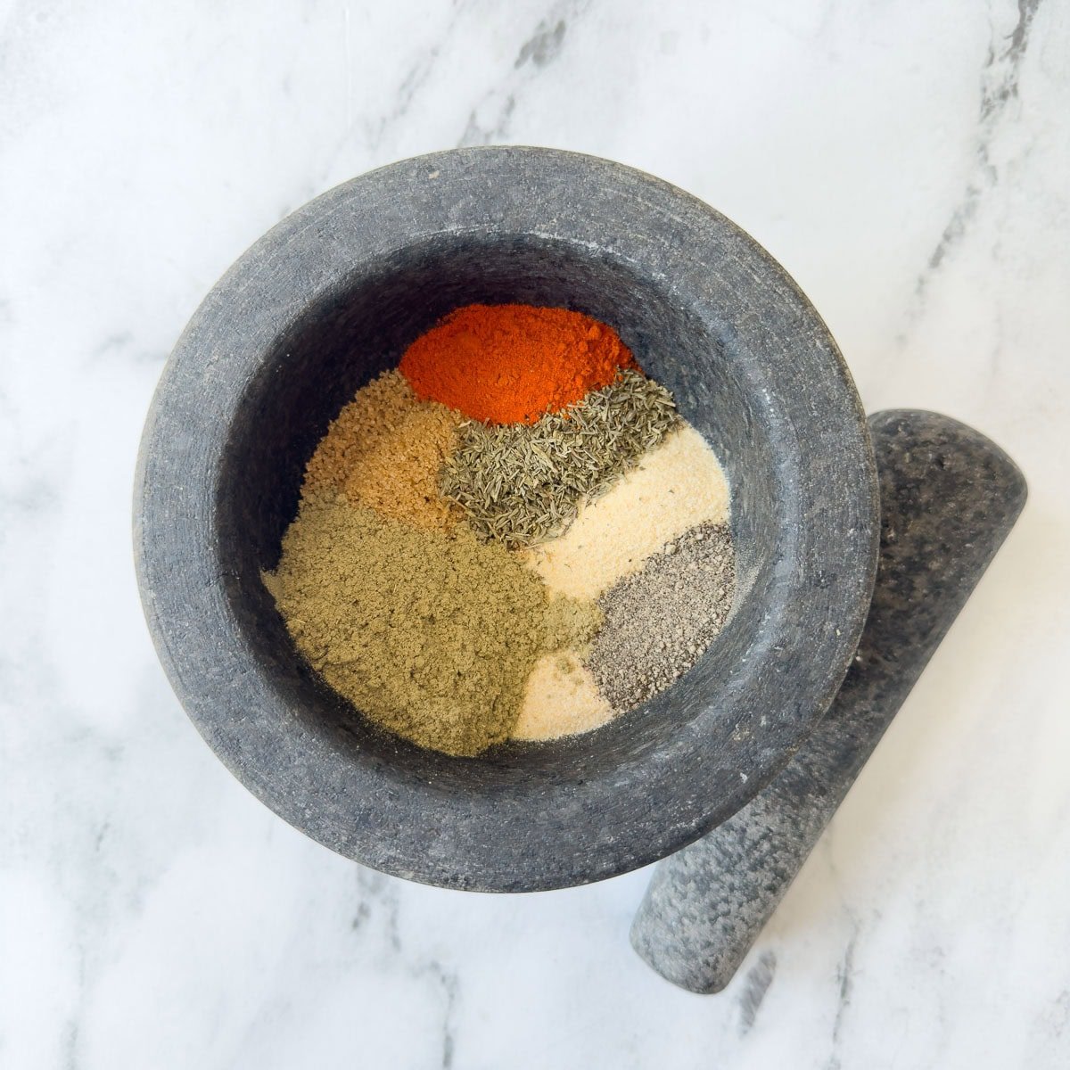 Brightly colored herbs and spices in the bowl of a gray mortar for blending.