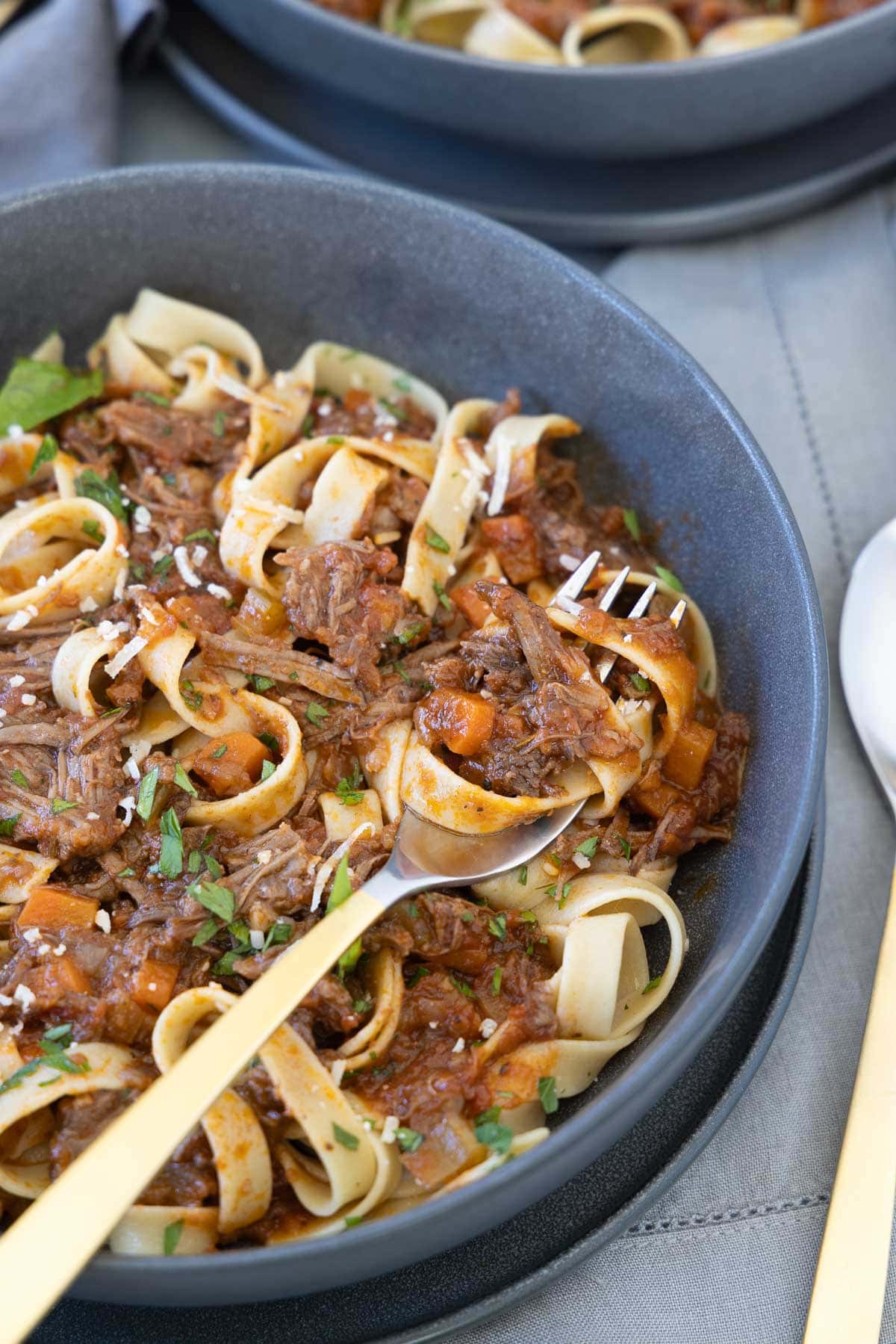 Twirled pasta on a fork of beef rib ragu in a gray bowl with carrots and parsley.