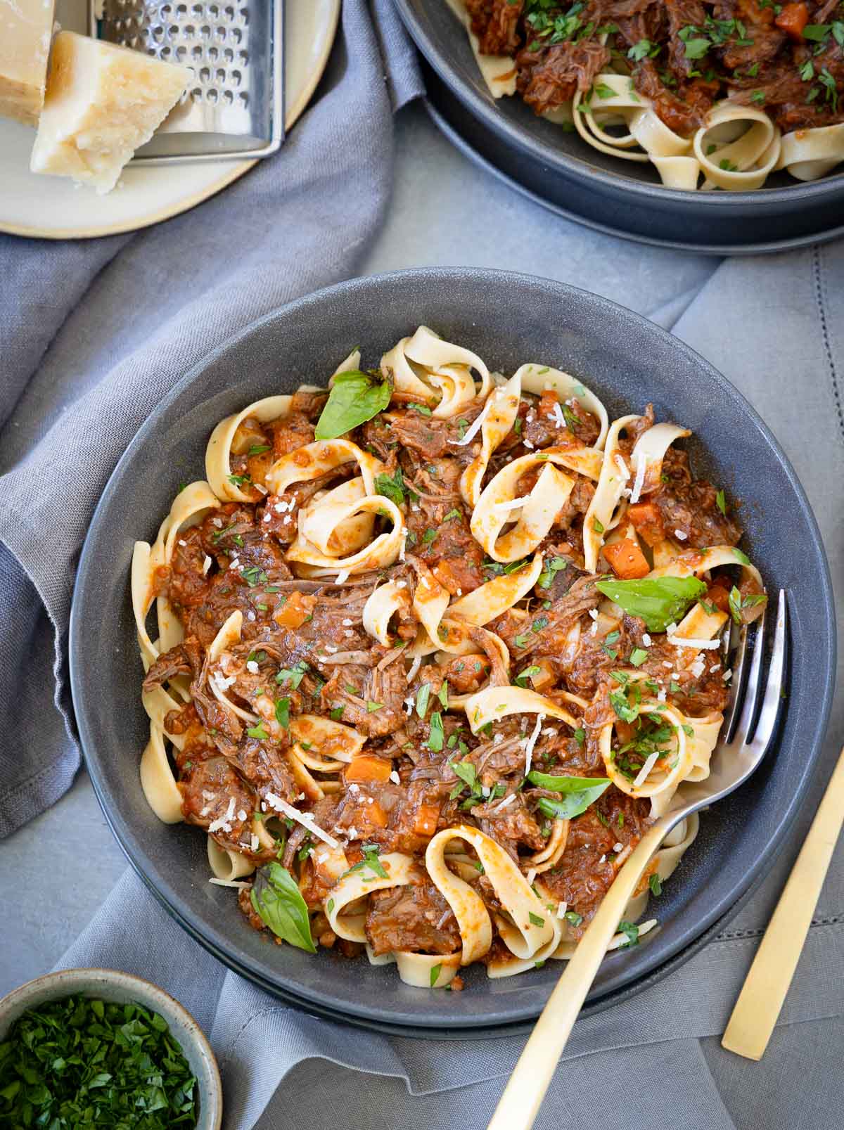 Gray bowl of wide pasta noodles with beef short rib ragu with carrots, and basil leaves.