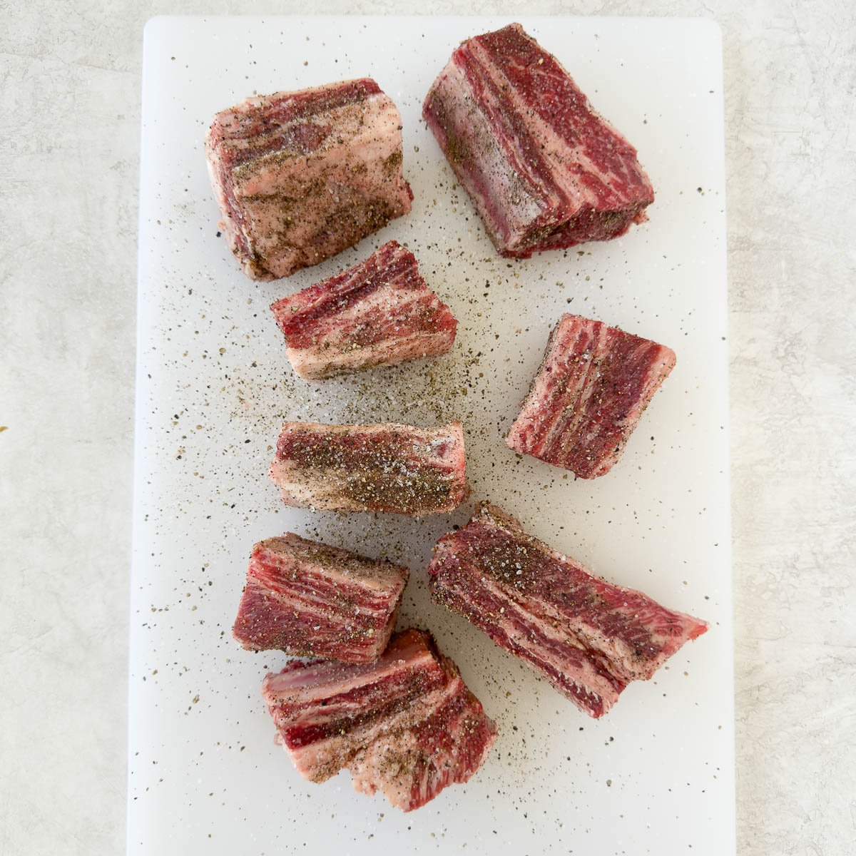 Salt and pepper seasoned beef short ribs on a white cutting board.