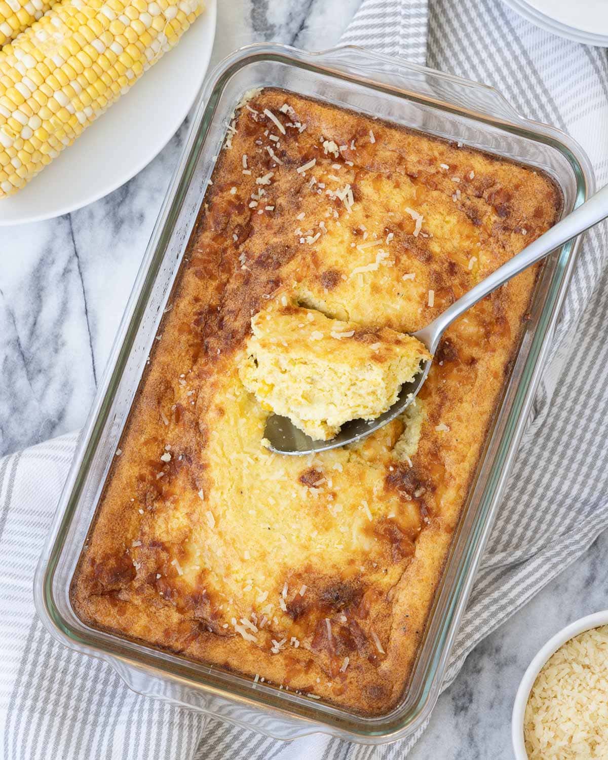 A golden corn souffle casserole in a glass baking dish with corn cobs and parmesan nearby.